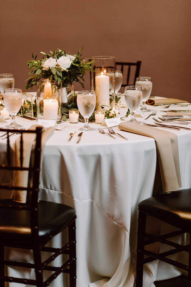 A reception table with white and beige linens, plus candles and greenery at the center