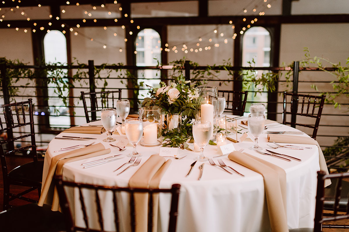 A reception table with white and beige linens, plus candles and greenery at the center at the Bell Tower in Nashville