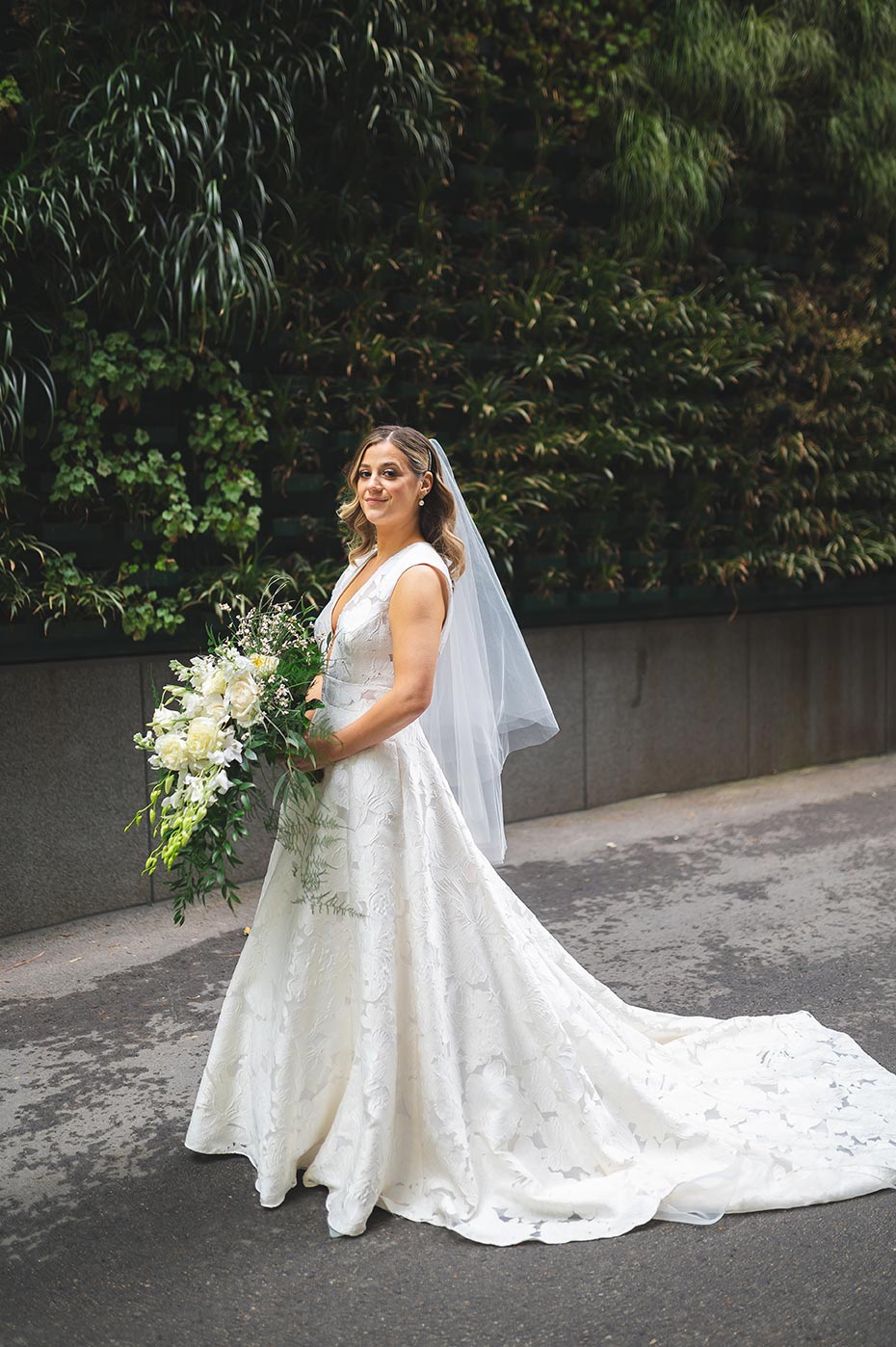 Bride poses in v neck lace detail gown holding a while bride holds a stunning, cascading bouquet full of wild greenery and bright white blooms.