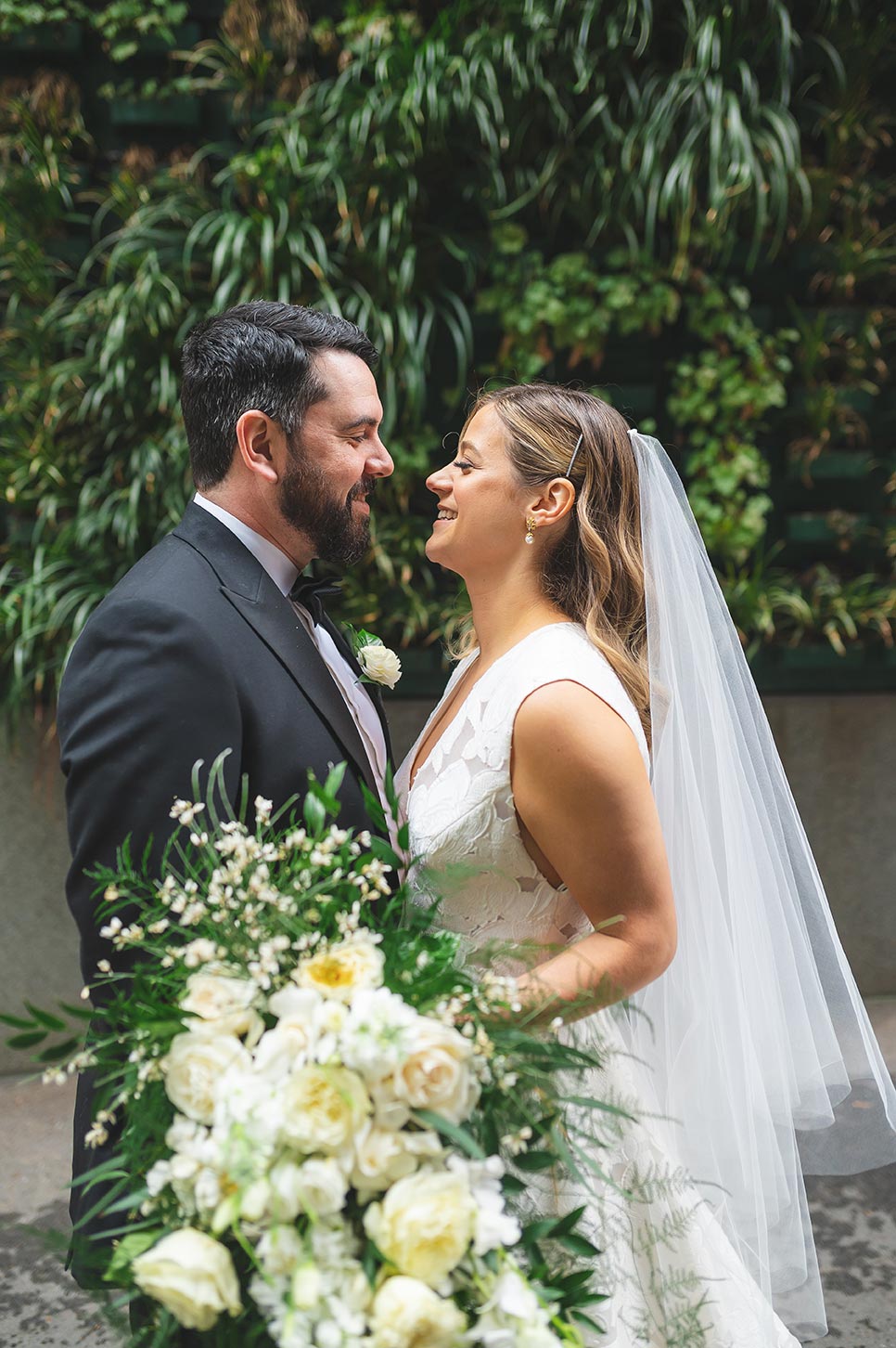 Bride and groom pose while bride holds a stunning, cascading bouquet full of wild greenery and bright white blooms.