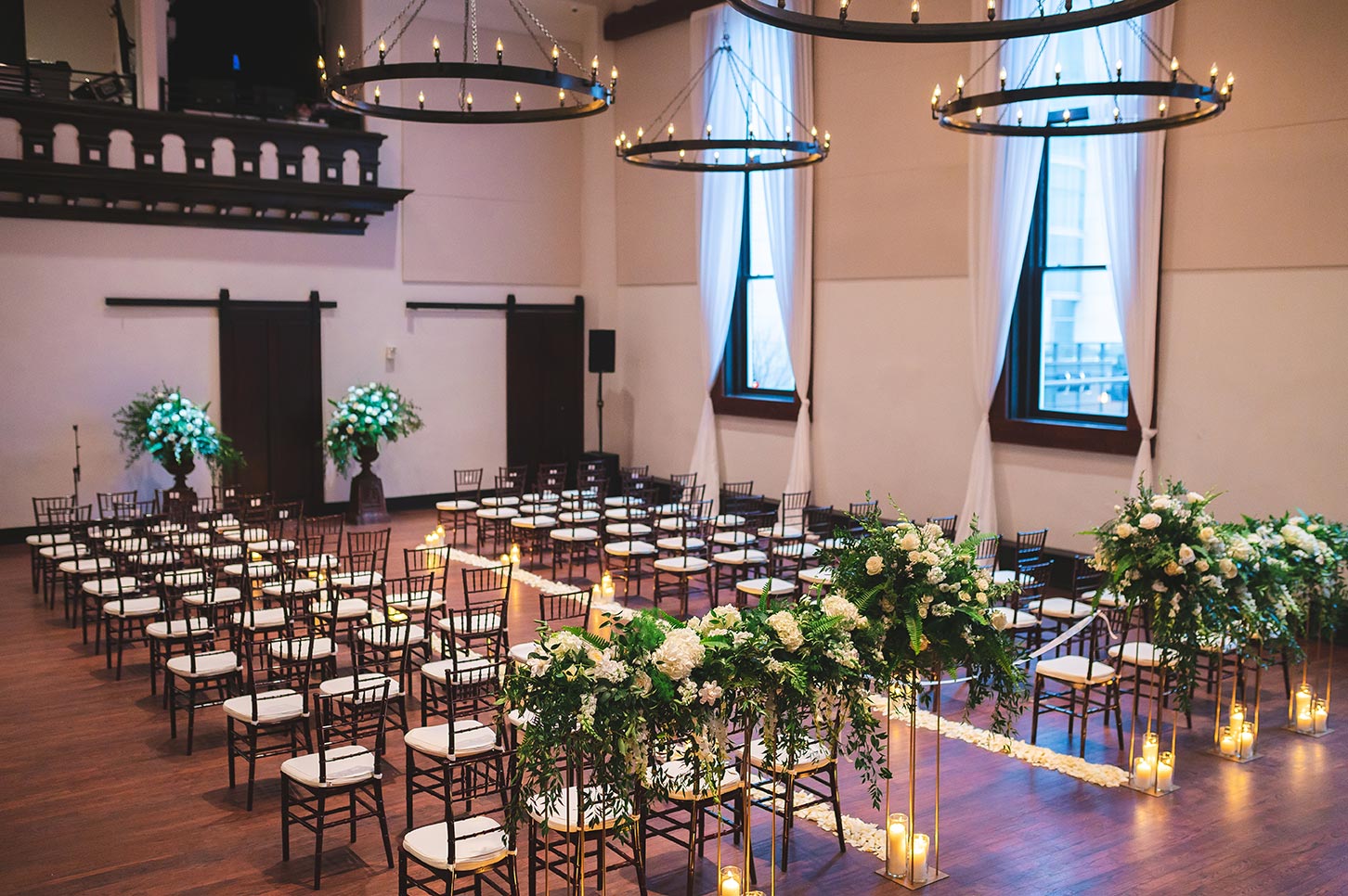 Ceremony space with large standing florals dark colored chairs rose petal lined aisle with candles