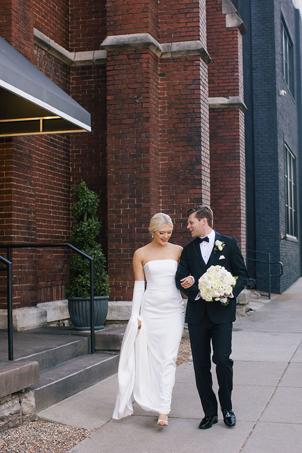 Classic Bride with Sleek Low Bun and Pearl Earrings in Column Gown with Hand-tied Traditional White Bouquet | Groom in Black Tuxedo Pose in Front of The Bell Tower in Downtown Nashville