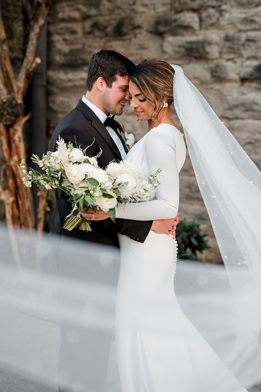 bride and groom embrace as veil with pearl details blows around them