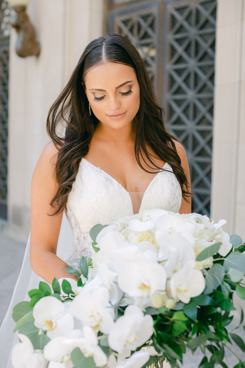 Bride in V-neck gown and long loose wavy hair looks down at white bouquet of orchids