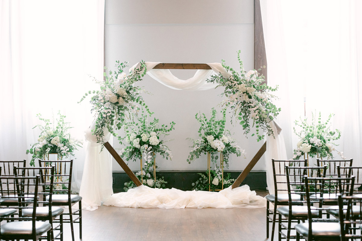 Wedding ceremony setup with hexagonal wooden arch decorated in white drapes and white floral arrangements with whimsical greenery sprigs