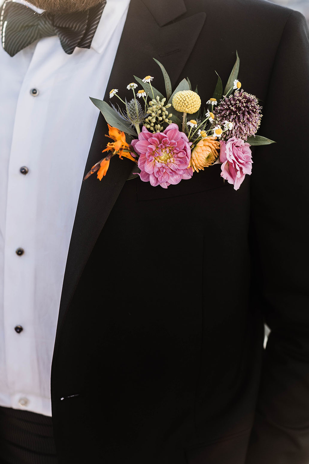 Close up of groom's bright and bold boutonniere with pink, orange, and yellow flowers against green leaves