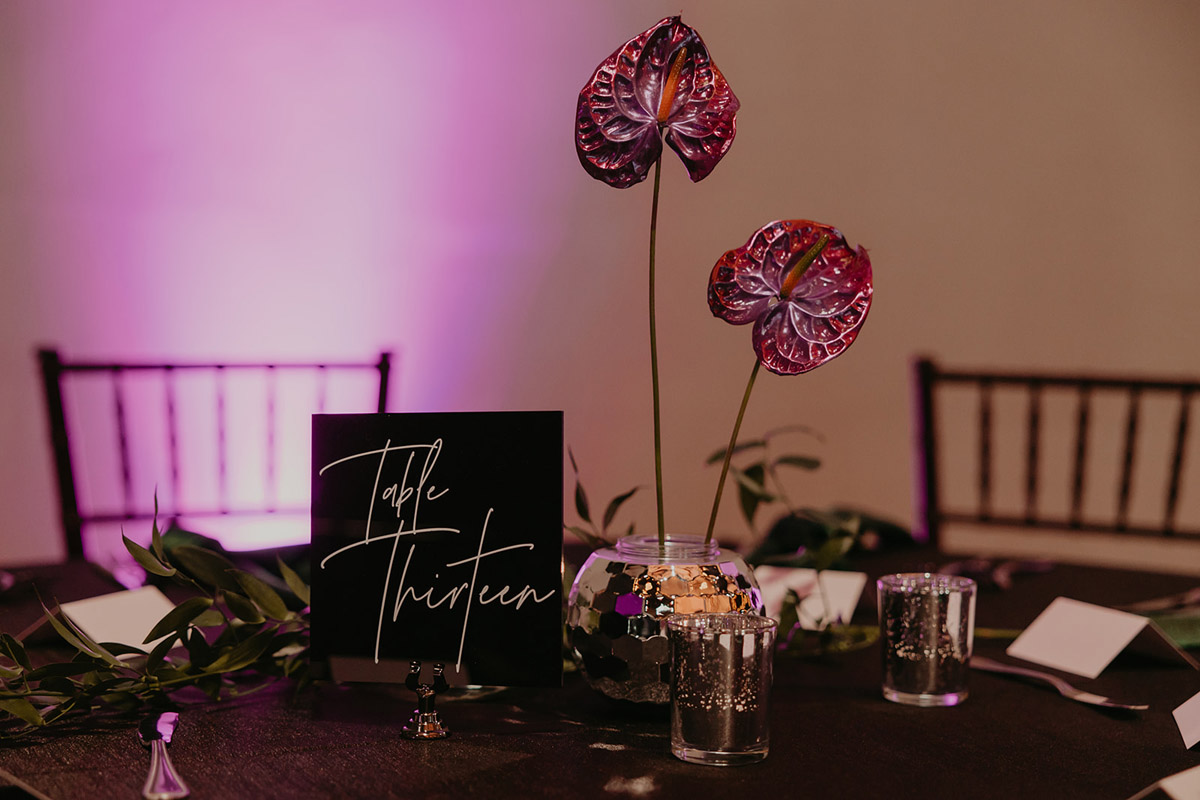 A wedidng table centerpiece with custom hand-painted anthuriums in disco ball bud vases with black acrylic table signs