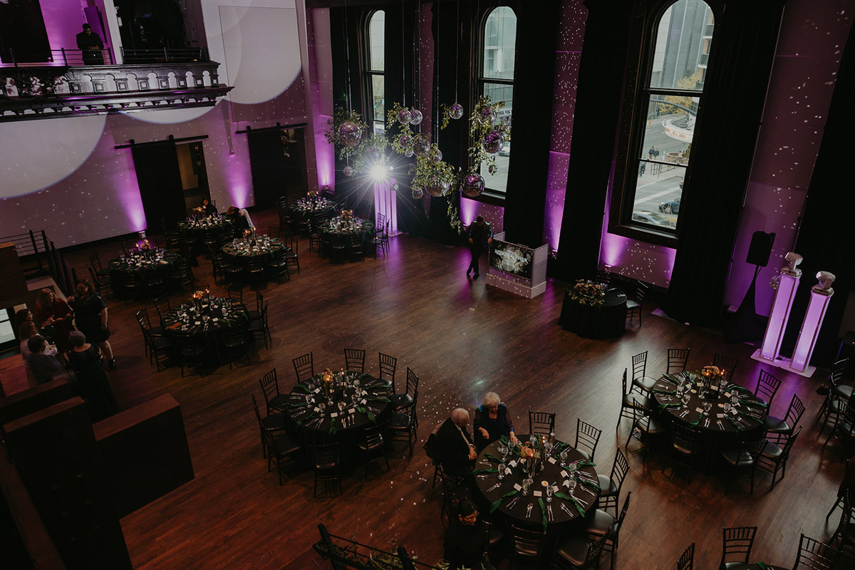 Wedding reception setup with disco ball installation hanging from the ceiling with greenery, tables decorated with black linens, and purple lighting