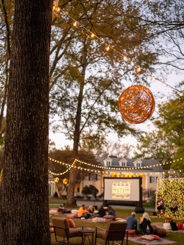 Outdoor movie night on a lawn with a big screen, lounge seating and string lights through the trees