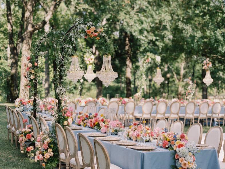 Garden reception designed with long blue tables, flower-covered arches and chandeliers suspended from the trees
