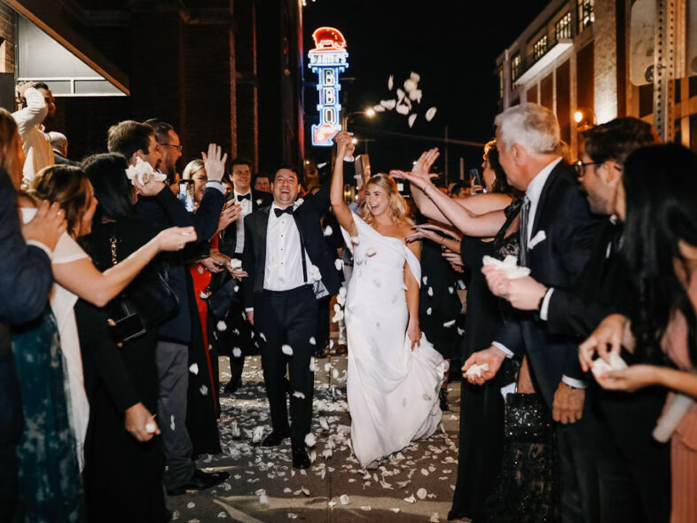 Newlyweds running through a crowd of cheering guests tossing white petals on a Nashville street at night