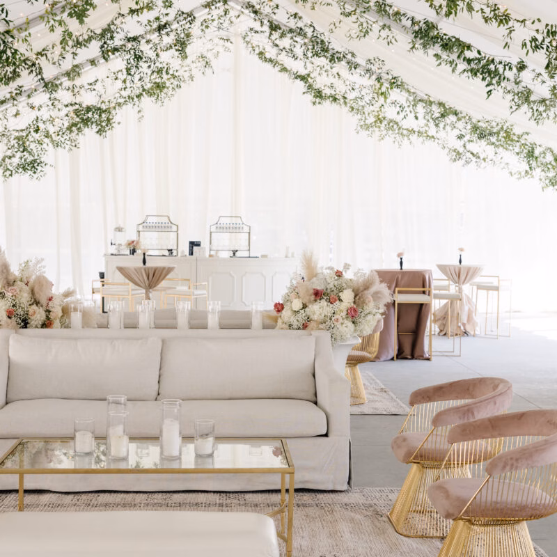 Bridal lounge in a white tent with cream sofas, blush accent chairs and greenery draped overhead