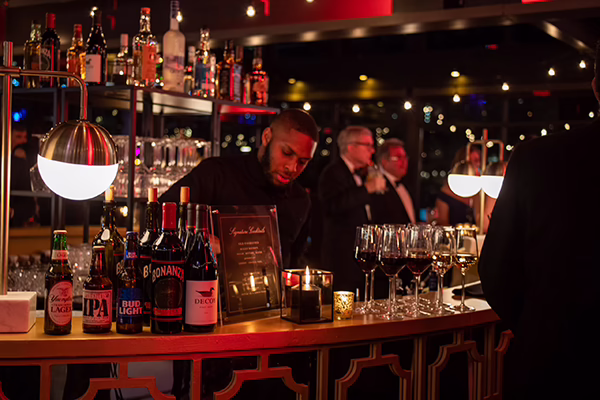 Bartender pouring drinks behind a lit bar stocked with wine, beer and spirits as guests wait