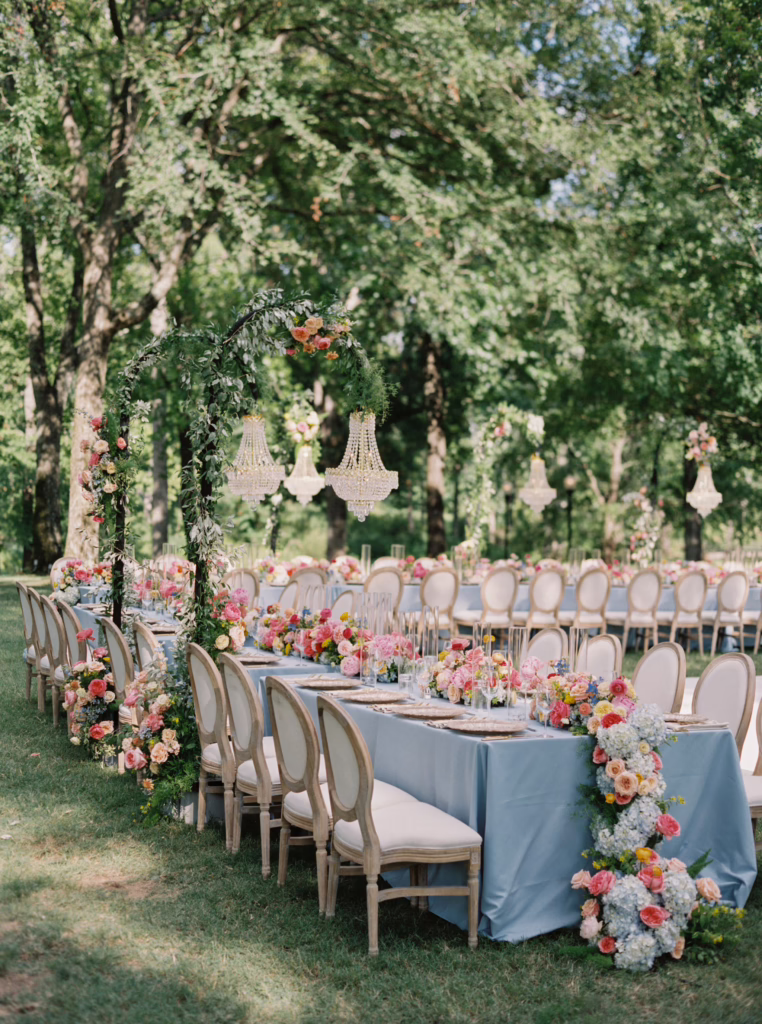 Long garden reception tables with blue linens and pastel flowers under trees hung with chandeliers
