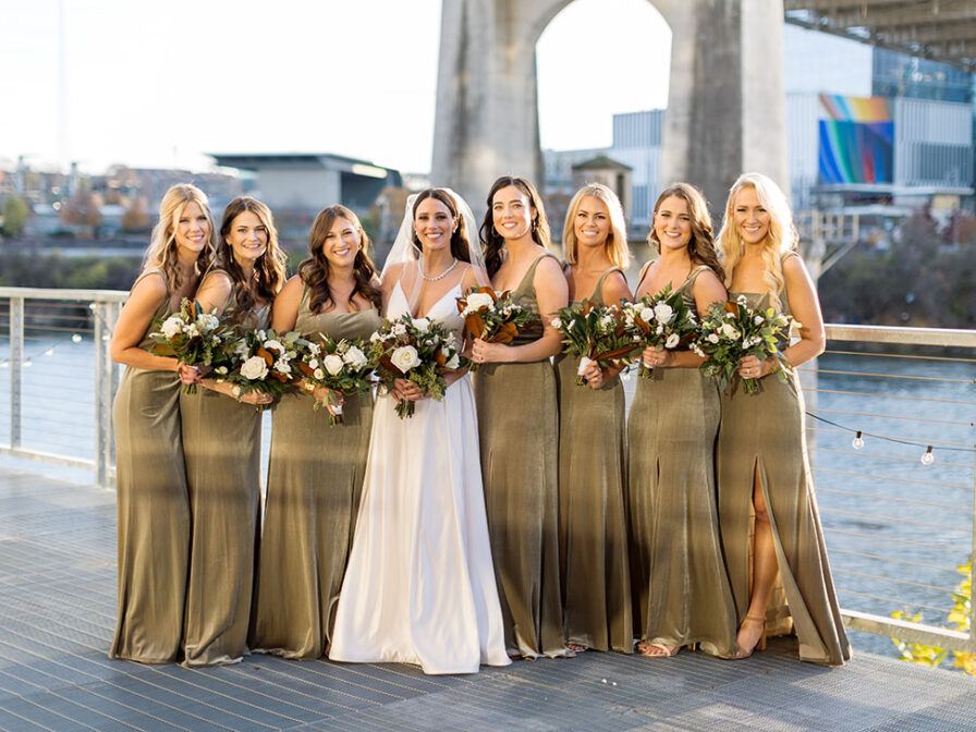 Bride posing with bridesmaids wearing green velvet bridesmaids dresses on the riverfront in downtown Nashville