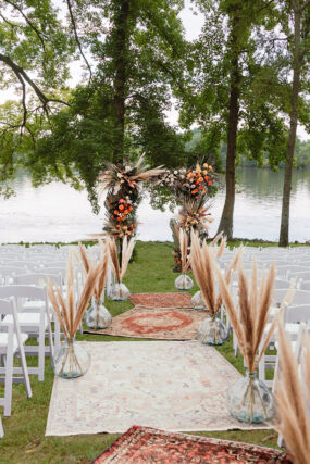 Bohemian wedding ceremony setup on the lakeside lawn with oriental rugs and pampas grass aisle markers