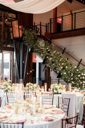 delicate gold details, lush floral arrangements in hues of white and pink with verdant greenery, the soft glow of candles, and white draping from the beams
