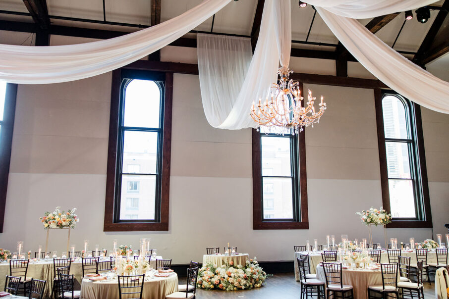 delicate gold details, lush floral arrangements in hues of white and pink with verdant greenery, the soft glow of candles, and white draping from the beams